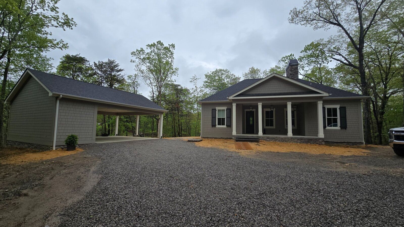House with carport on gravel driveway in a wooded area under a cloudy sky.