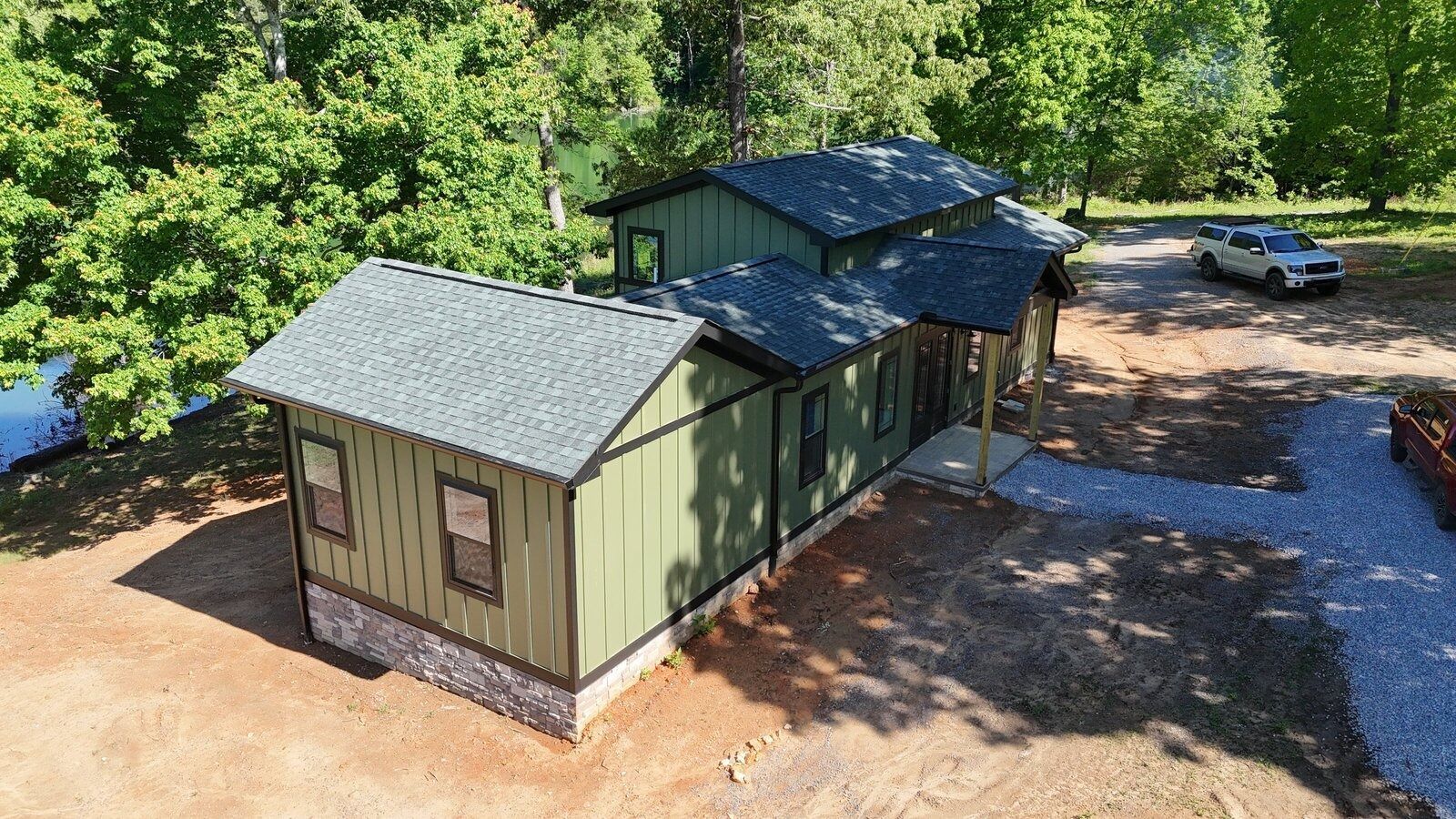 Green house with dark roof and gravel driveway; car parked nearby, trees in background.