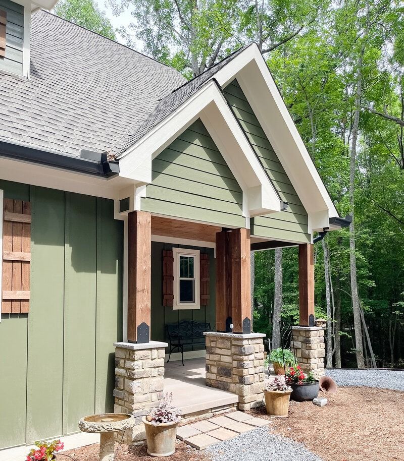 Green cabin with stone columns, porch, and a gray shingled roof, set amongst trees.