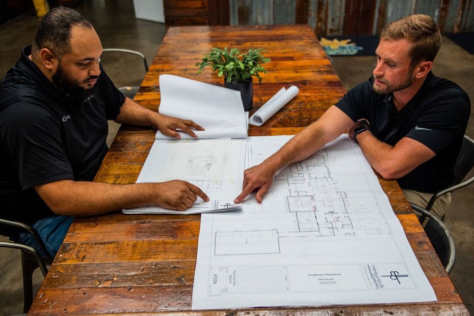 Two men review blueprints at a wooden table, discussing a project.