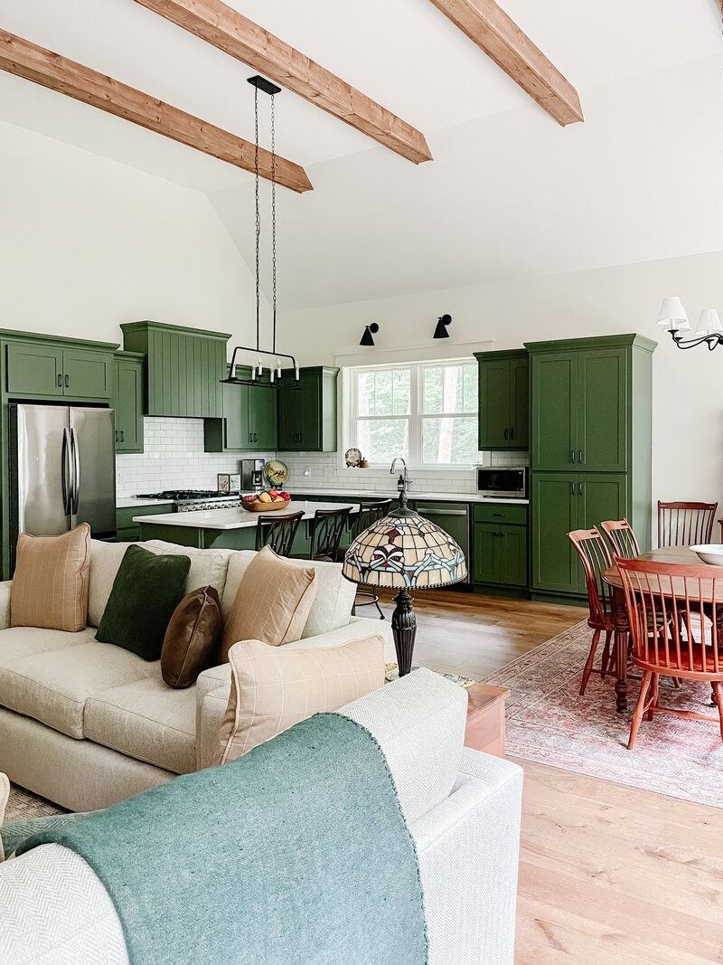 Green kitchen with white countertops and cream-colored sofa. Wooden beams, round table, and a patterned lamp.