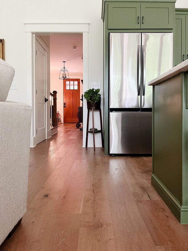 Light wooden floor in kitchen, leading to hallway with open door. Olive green cabinets and stainless steel refrigerator.