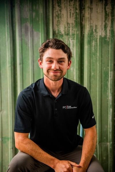 Man with a slight smile wearing a black polo shirt, sitting in front of a green wall.