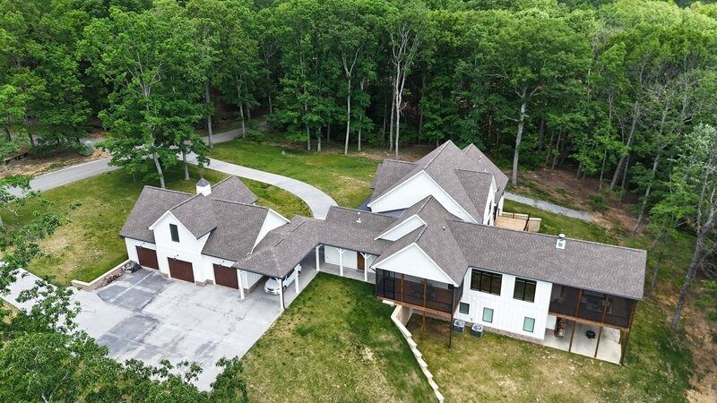 Aerial view of a white house with a brown roof and a long driveway surrounded by green trees.