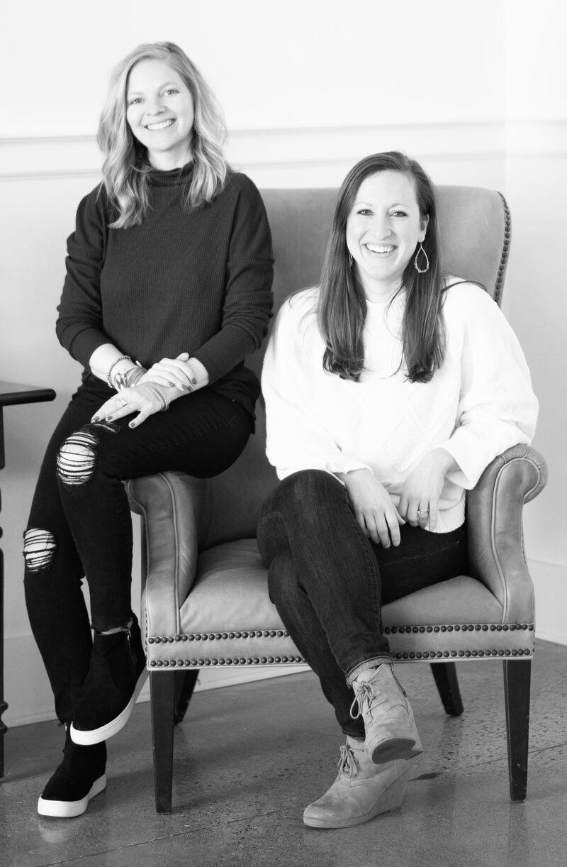 Two smiling women pose; one sits in a chair, the other beside her. Black and white photo.