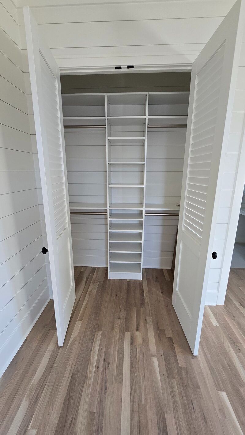 A walk-in closet with white shelves, rods, and doors.  Light wood-tone floors.