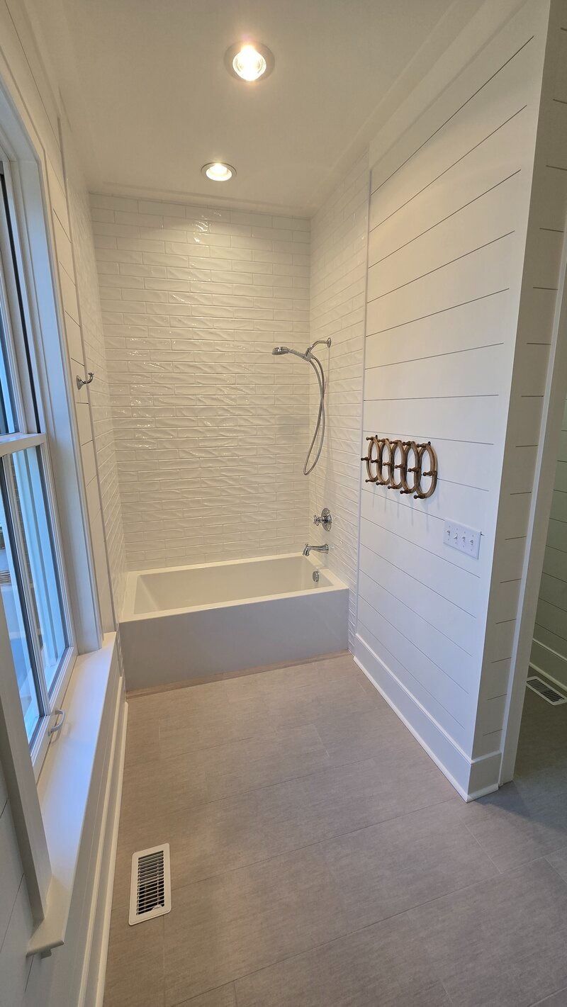 Small bathroom with white tiled wall, bathtub, and light-colored flooring; a window is on the left.