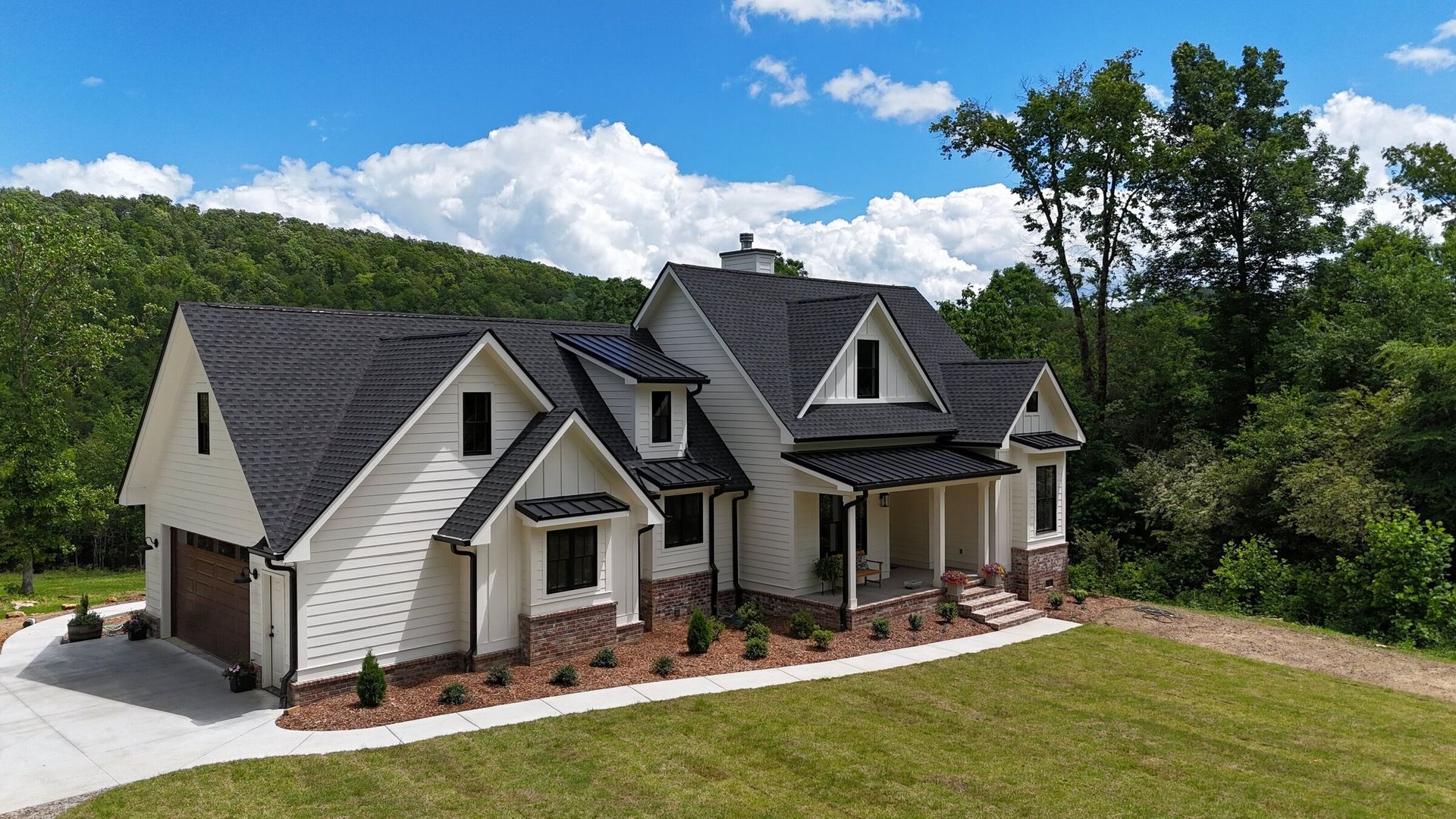 White house with black roof, brick accents, and green lawn against a forested hillside under a blue sky.