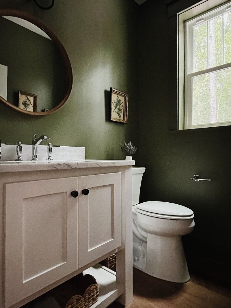 Green-walled bathroom with white vanity, toilet, round mirror, and a small window with light shining through.