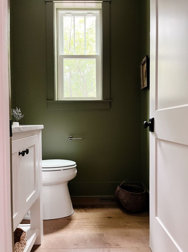 Small bathroom with olive-green walls, a white toilet, and a window. Ajar white door.
