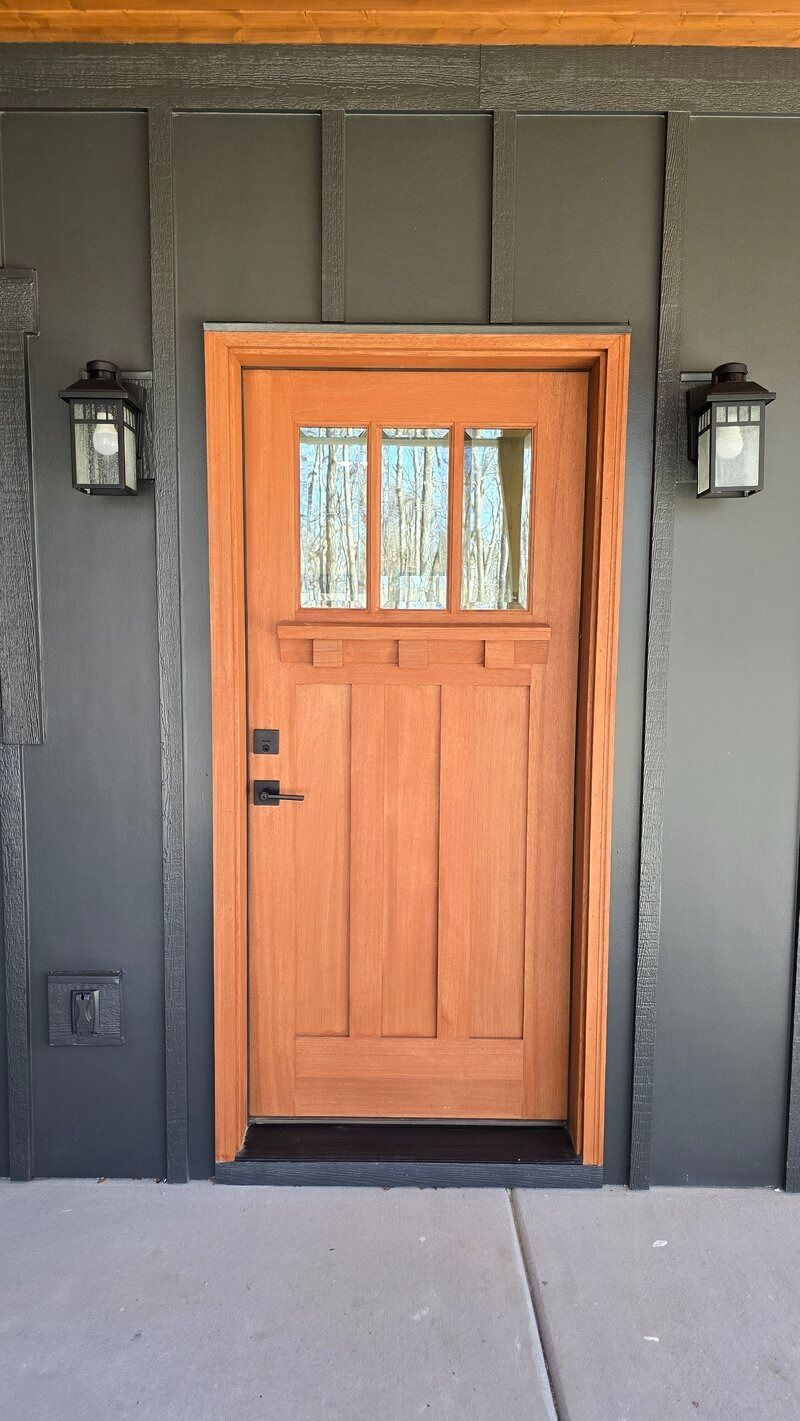 Wooden Craftsman-style front door with glass panels. Flanked by black wall lanterns, set in a dark gray exterior.
