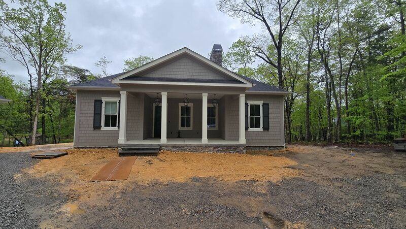 Tan house with gray shutters and porch, set in a wooded area with gravel and dirt.