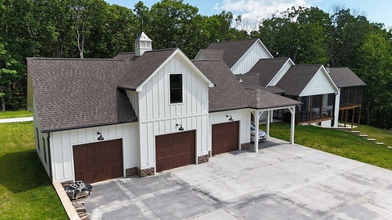 White farmhouse with brown roof, three-car garage, and attached carport. Green trees in the background.