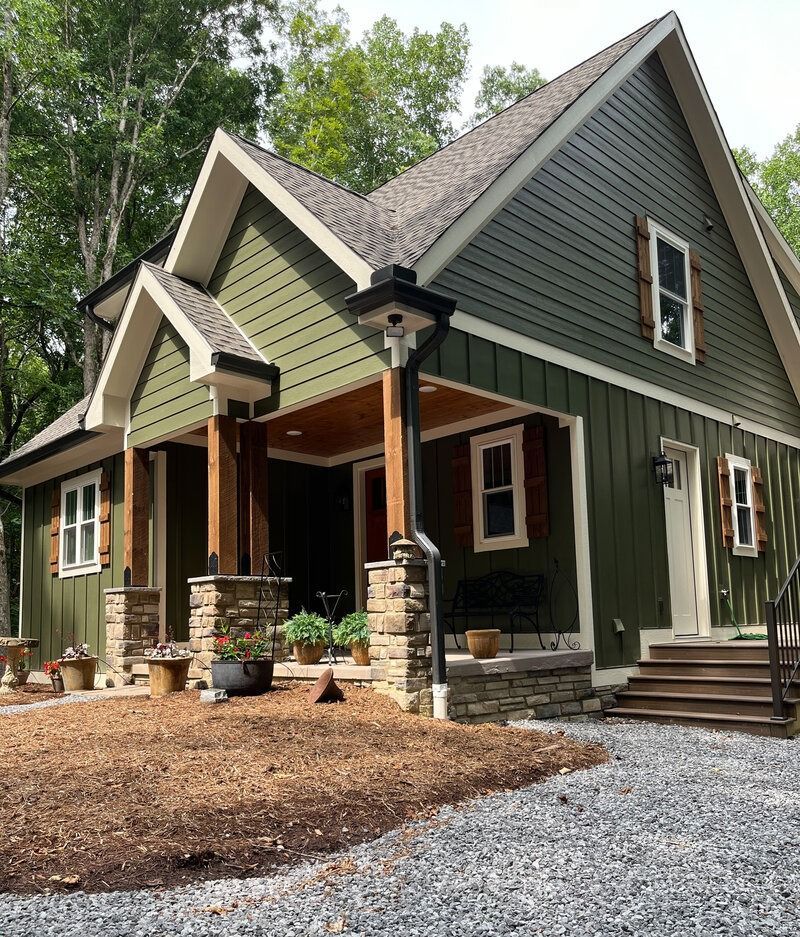 Green house with stone columns, brown shutters, and gravel path.