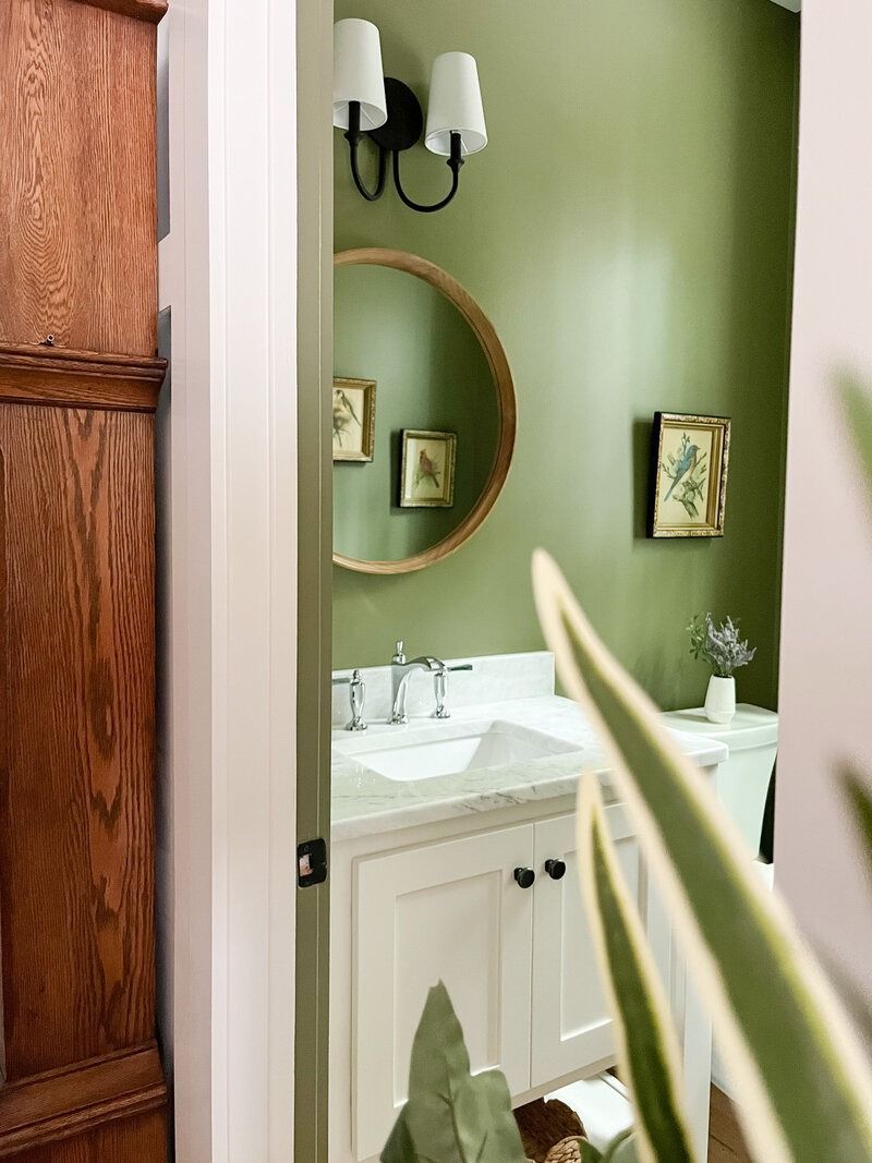 Bathroom with olive green walls, a white vanity, round mirror, and framed art. Wooden door frame on left.