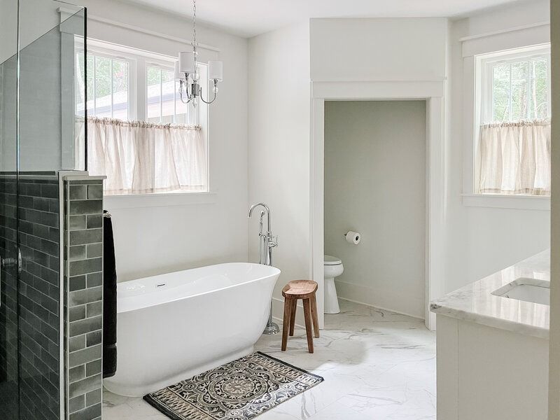 White bathroom with a freestanding tub, toilet, & sink; marble tile flooring, a shower with dark green tile, and two windows.