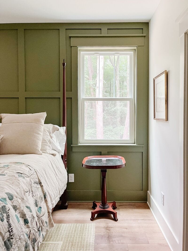 Bedroom with olive green paneled wall and window. Bed with patterned quilt, small table.