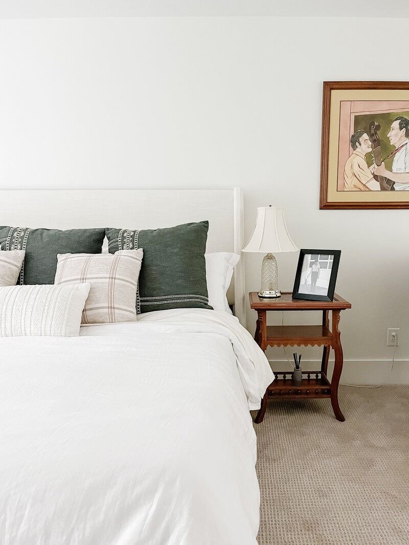 Bedroom with white bed, dark green and white pillows, wooden nightstand, art.