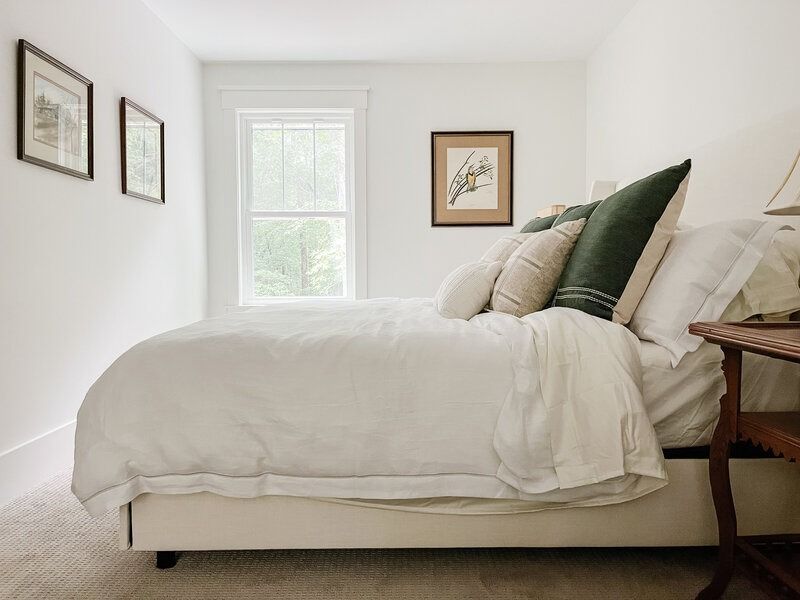 Bedroom with white walls, bed, window, and framed art. Beige carpet, decorative pillows, and a wooden side table.