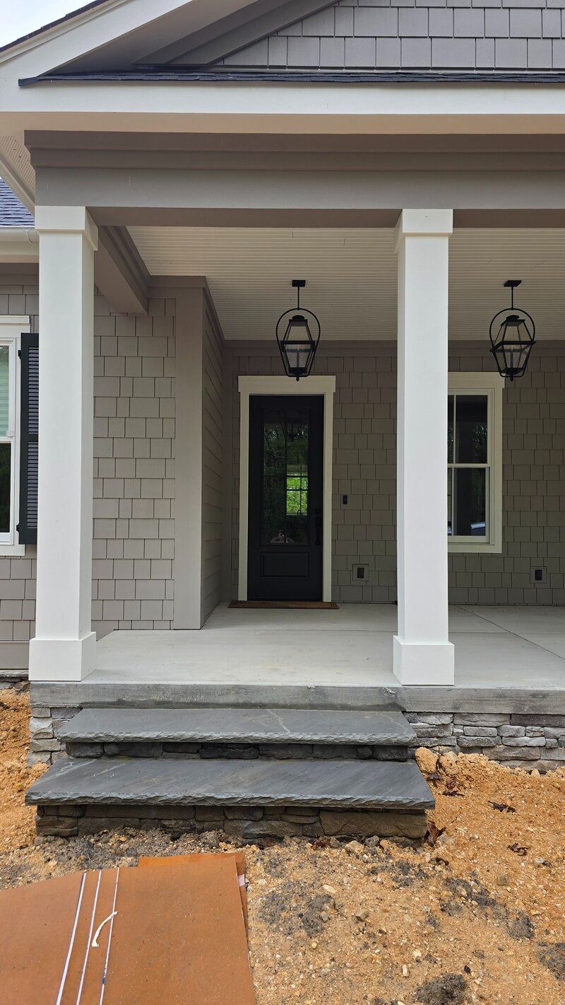 Front porch with gray shingle siding, white columns, black door, and stone steps.
