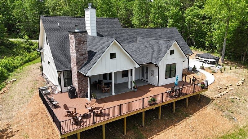 White farmhouse with brown deck, chimney, and dark roof in a wooded area.