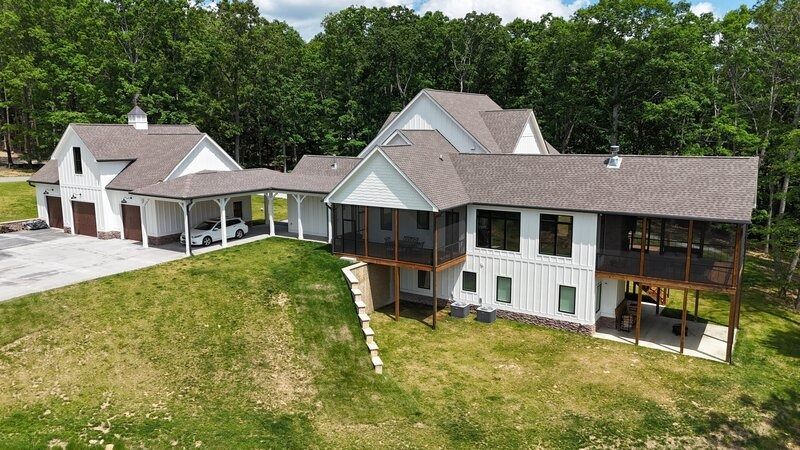 White farmhouse with attached garage, carport, and screened porch on a grassy hill, surrounded by trees.