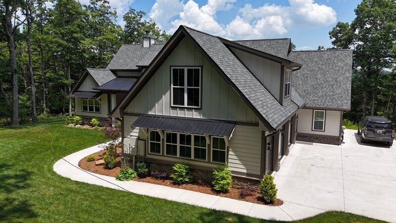 Two-story house with gray roof & green siding. Garage, windows, & a car are visible. Green lawn and trees surround the house.