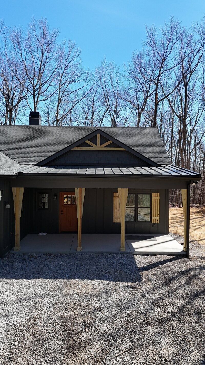 A cabin with a porch and wooden supports, a grey roof, and a gravel driveway, against a backdrop of trees.