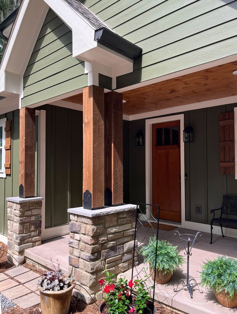Green house entrance with stone pillars, wooden beams, brown door, and potted plants.