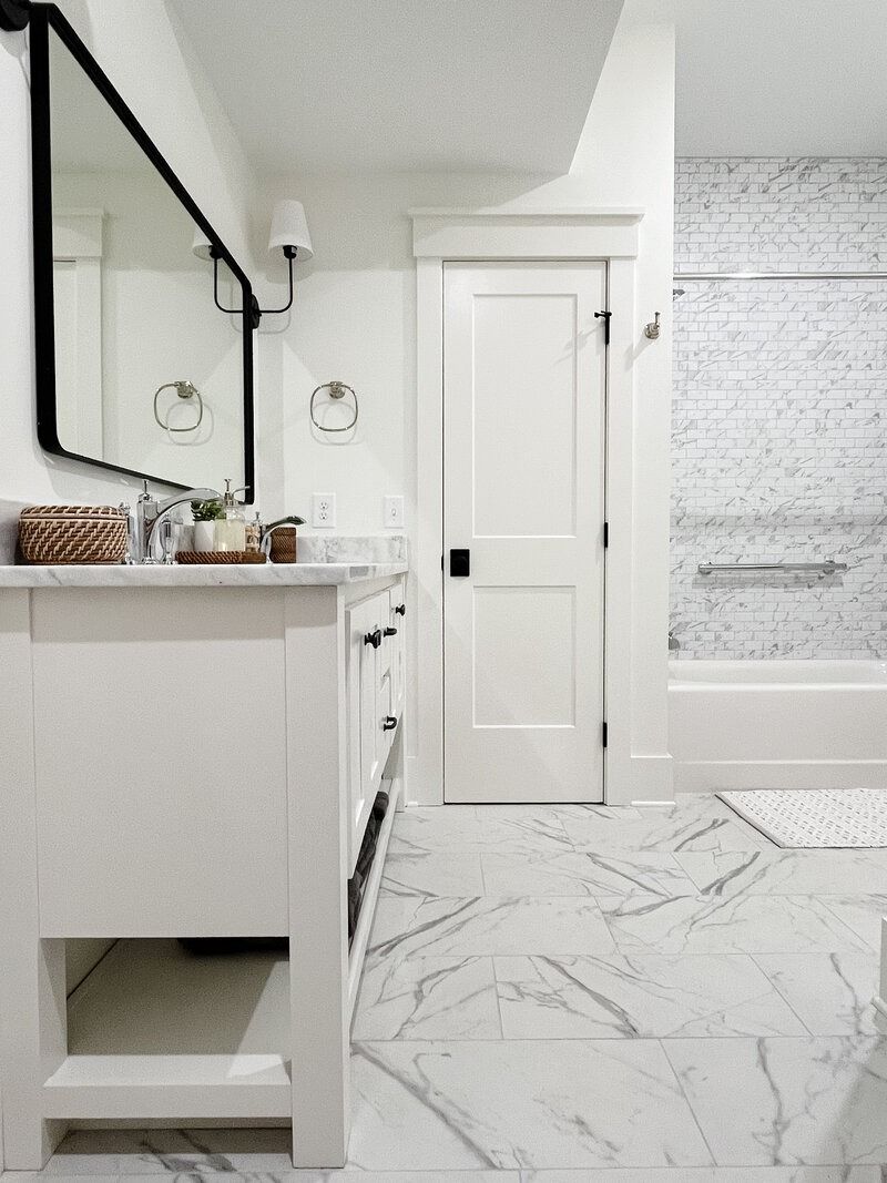 White bathroom with marble floor, vanity, door, and shower. Black accents include mirror frame, sconce, and hardware.