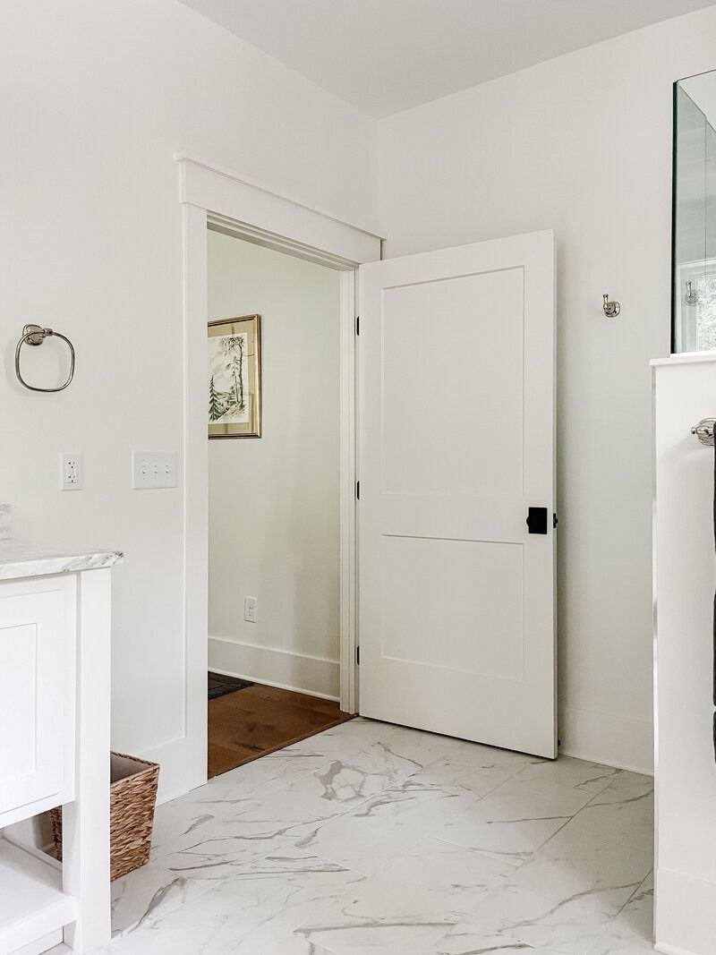 White bathroom with an open door, leading to a hallway, and marble-look flooring.