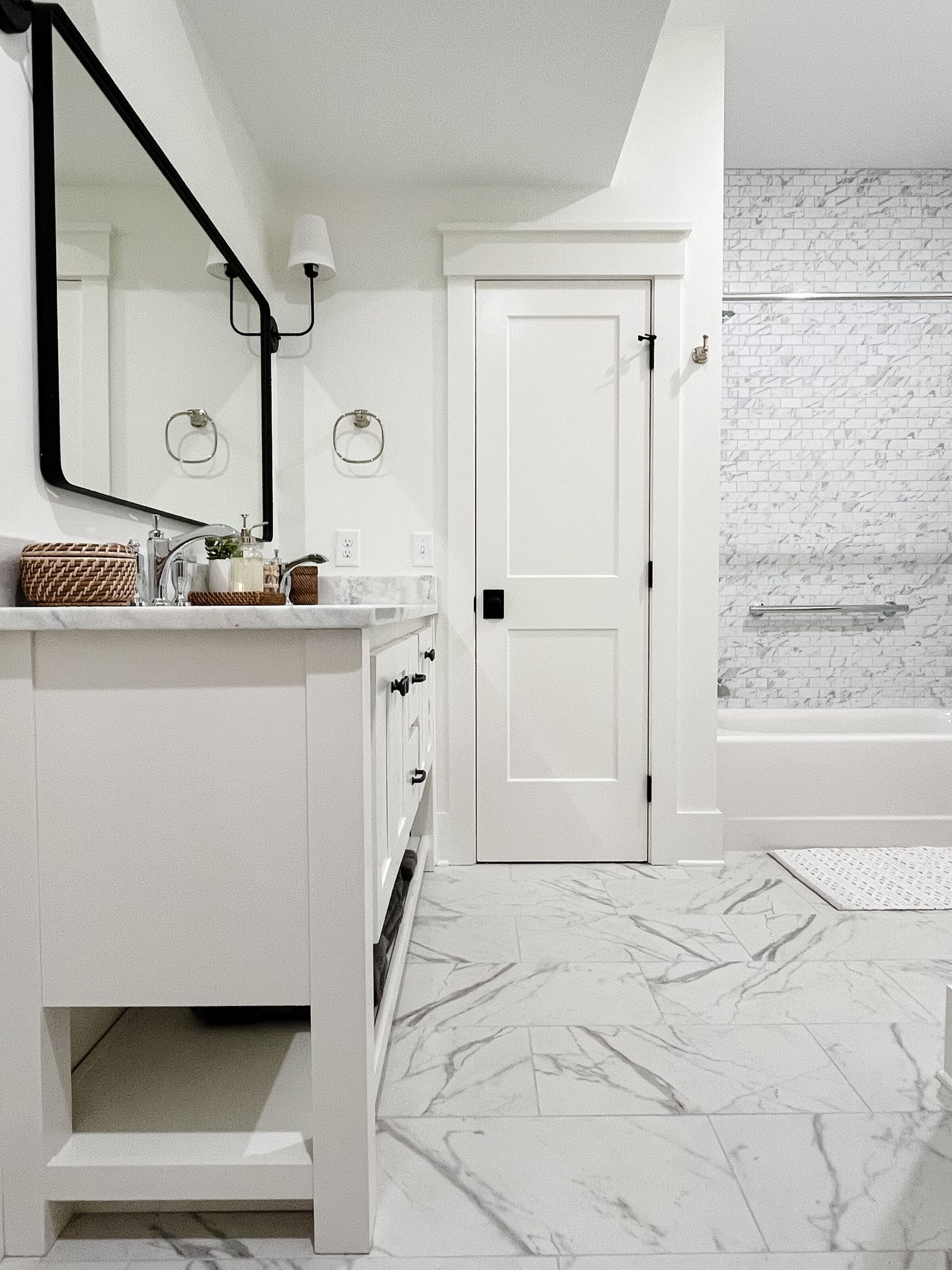 White bathroom with marble floors, vanity, and tub. Black-framed mirror and door.