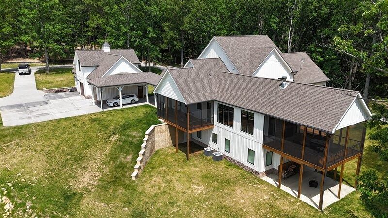 White house with a screened porch and attached carport on a grassy hill; driveway and trees in the background.