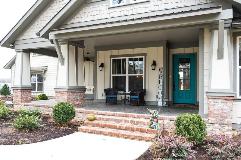 A home's covered front porch with a teal door, brick steps, and small bushes.