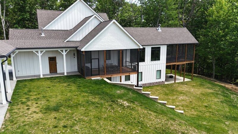 White house with brown roof and screened porch on a grassy hill, surrounded by trees.