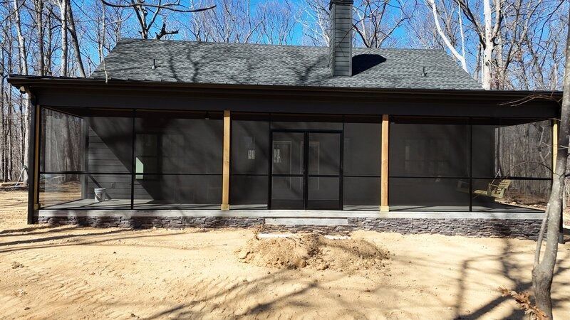 Screened-in porch on a building with a dark roof and chimney, set in a wooded area on a sunny day.