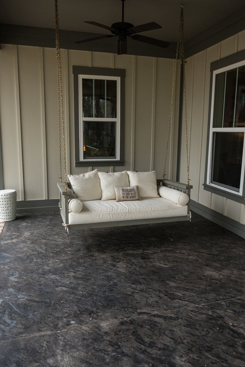 Covered porch with swing, two windows, and dark flooring. The swing has white cushions and pillows.