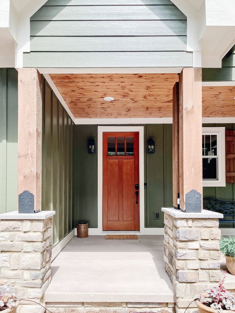 Entryway of a house with a red door, stone columns, wooden ceiling, and green siding.