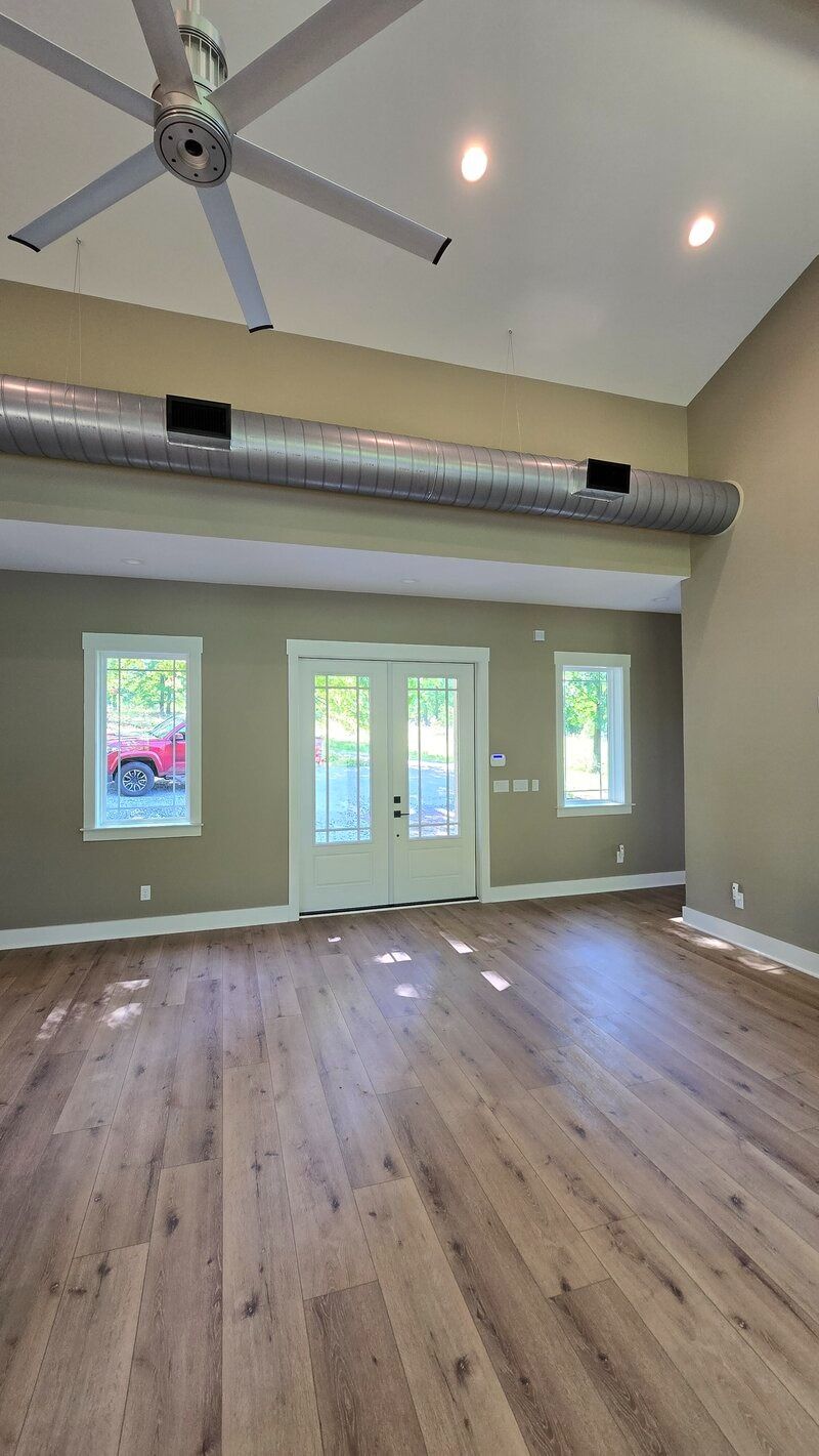 Spacious room with wood flooring, three windows, and double doors; exposed ductwork and ceiling fan.
