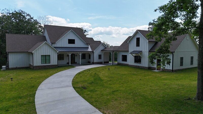 White farmhouse with brown roof and long, curved concrete driveway on green lawn.