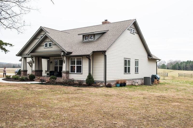 Beige house with a porch, brown roof, and brick foundation, set in a grassy field on an overcast day.