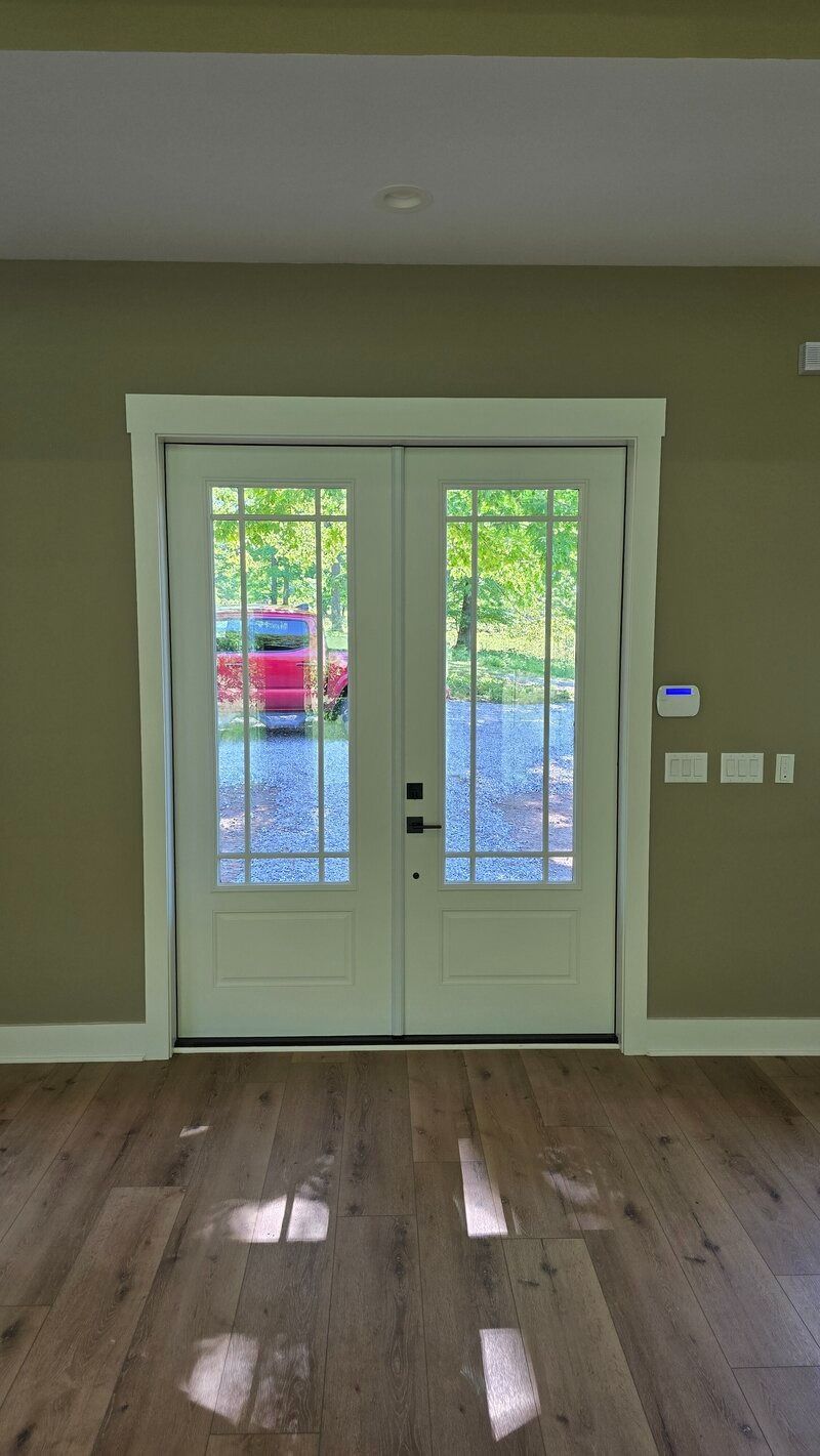 White French doors with multiple panes, framed in white, open to an outdoor view. Interior with wood floor, olive walls.