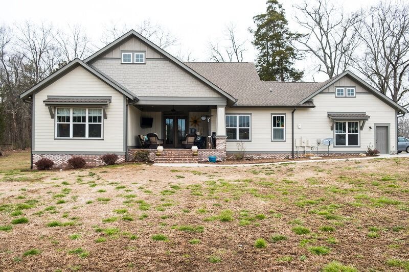 Tan house with gray roof, brick base, and front porch, surrounded by dry grass and bare trees.