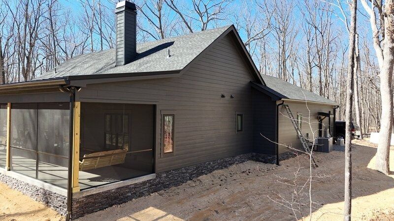 Dark-colored house with screened porch, chimney, and surrounding trees. Located on a dirt path.