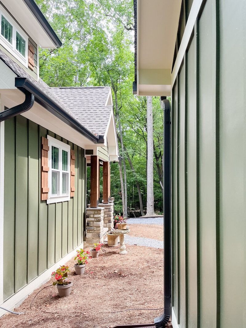 Two buildings with vertical green siding and brown roof line, path between, trees in background.