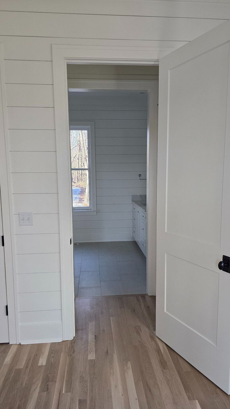 White interior doorway, leading to a bathroom. Shiplap walls, hardwood floors, and a white door are visible.