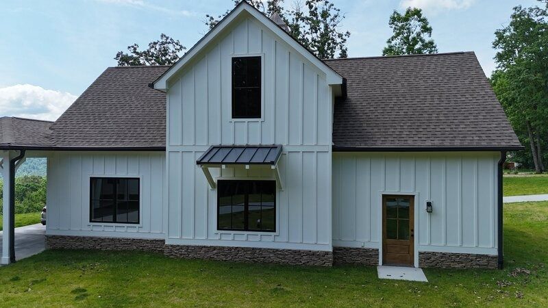 White farmhouse with dark trim, brown roof, and stone base on a grassy lawn.