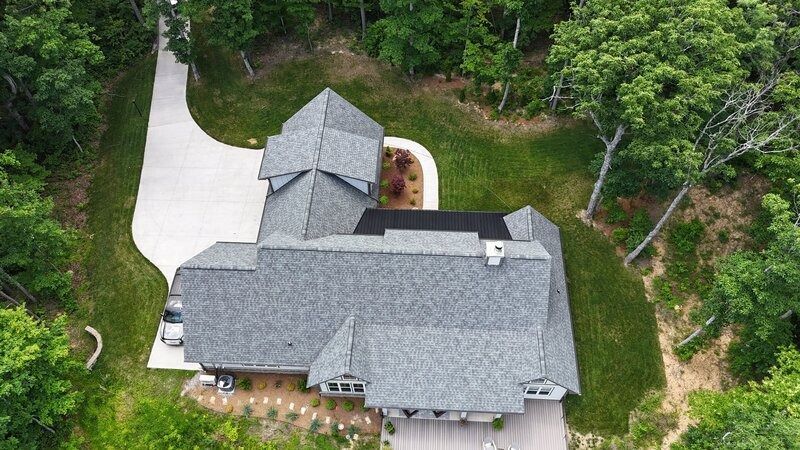 Aerial view of a gray-roofed house with a long driveway surrounded by green trees.