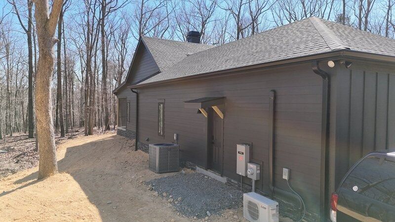 Dark-sided house with a black roof, surrounded by trees and a gravel driveway.