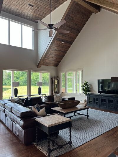 Spacious living room with high wood-paneled ceiling, large windows, dark brown leather sofa, and TV on a cabinet.